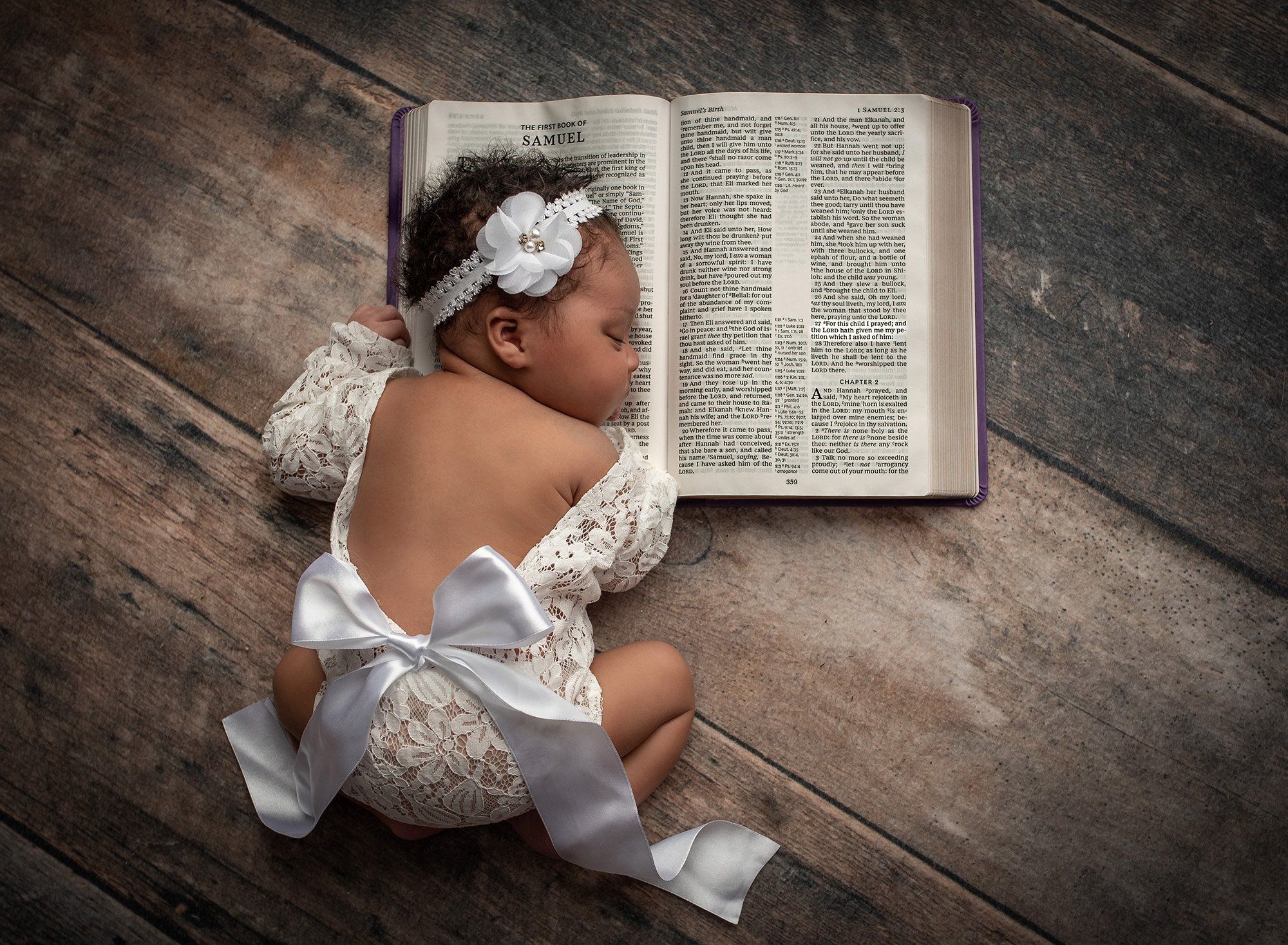 newborn baby girl wearing white laced dress with bow resting her head on the Bible