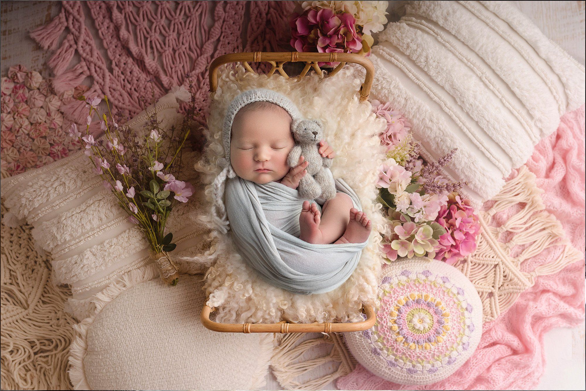 newborn baby girl sound asleep in grey bonnet on miniature rattan bed surrounded by soft pillows and macrame in white and pink