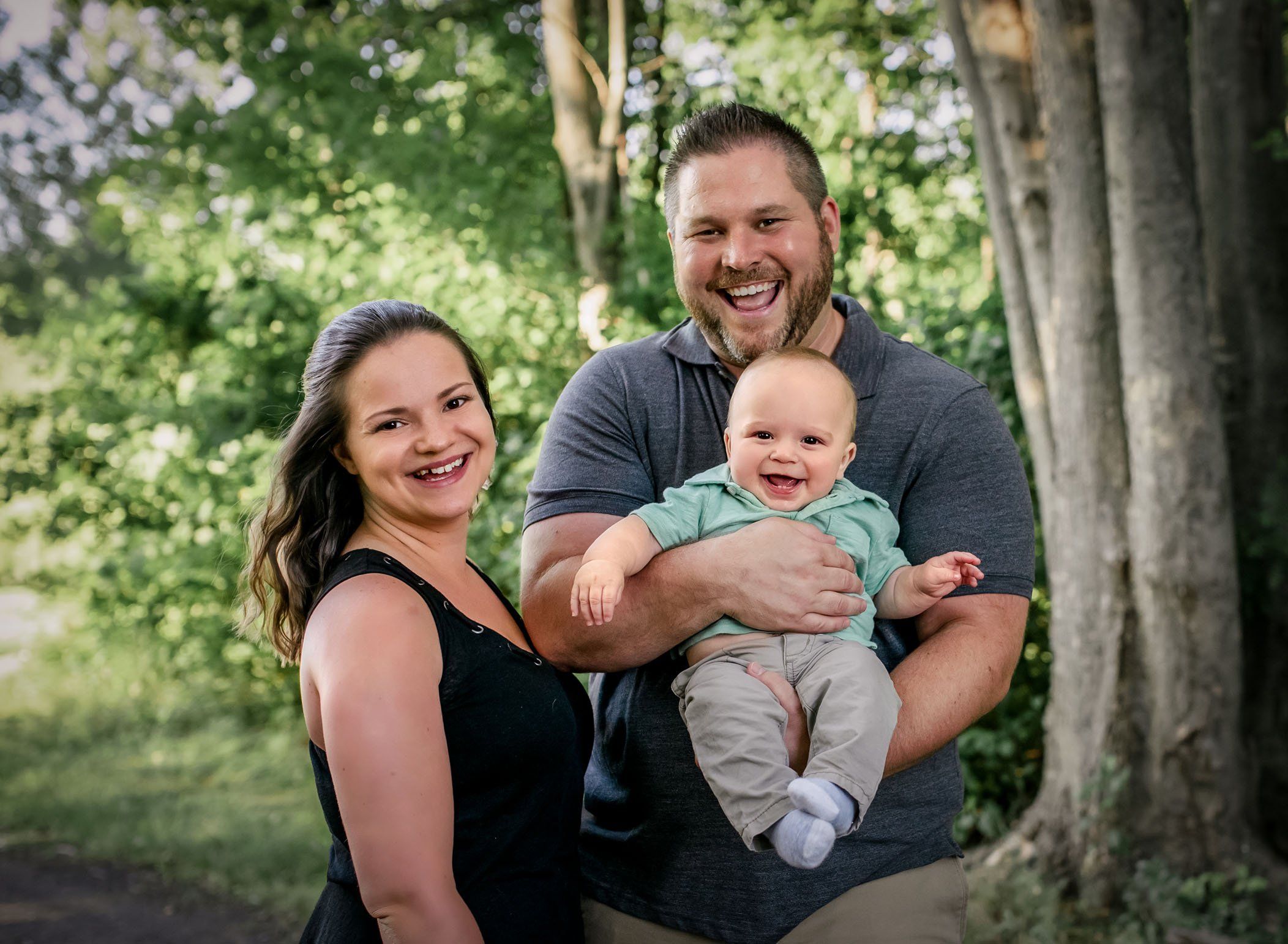 Dad holding 6 month old smiling baby boy in his arms with mom standing in close too