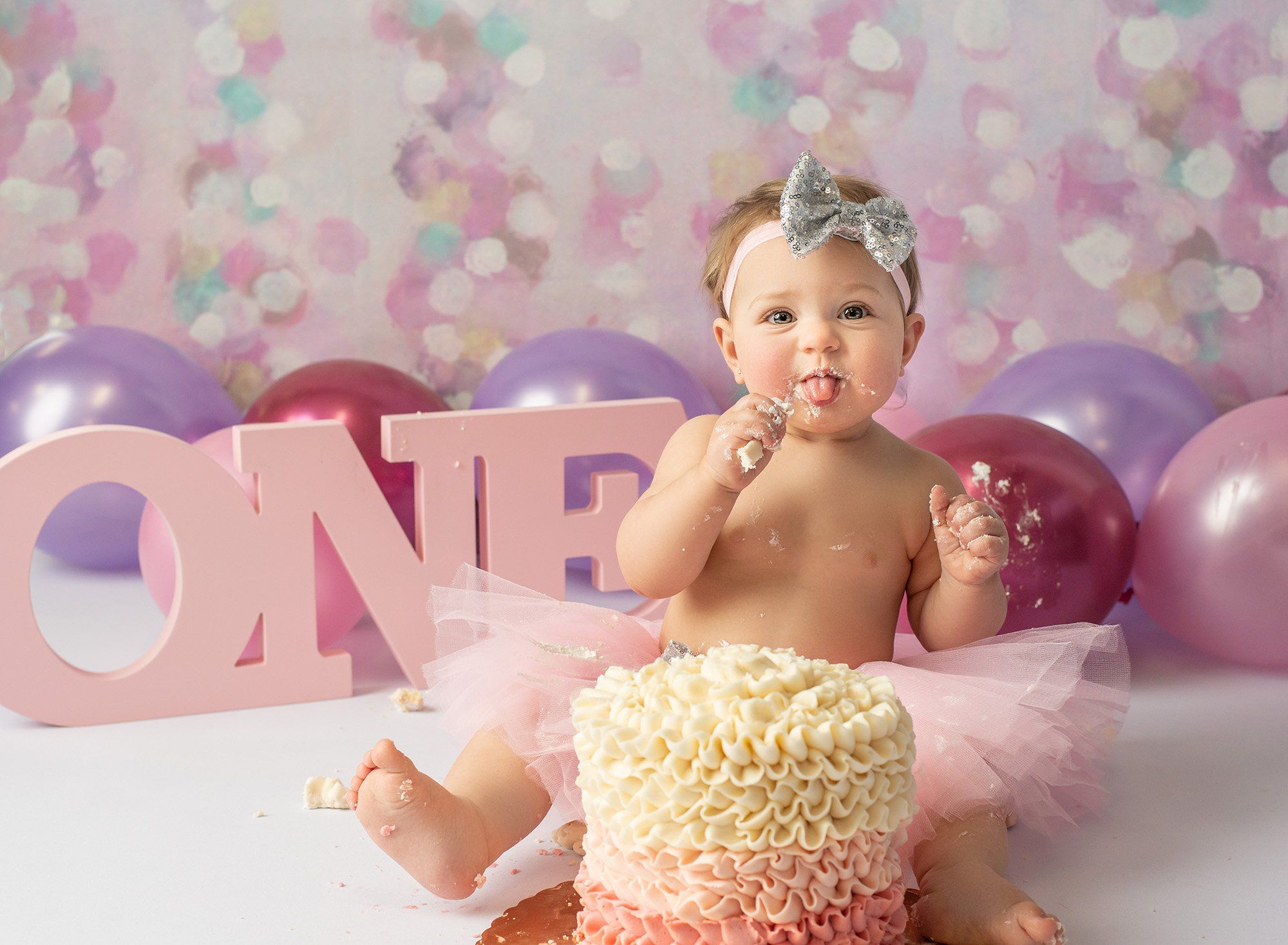 baby girl sticking out tongue at her one year cake smash surrounded by pink and purple balloons with an ombre pink cake