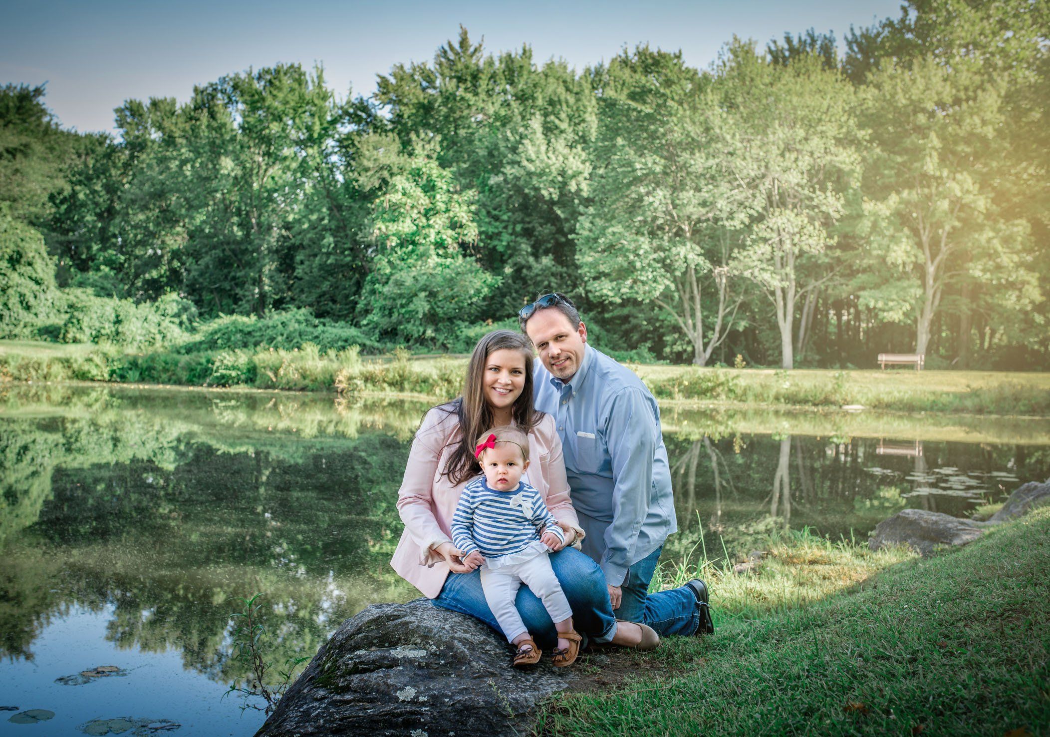 Mom, Dad and one year old baby girl sitting in front of pond in summer garden