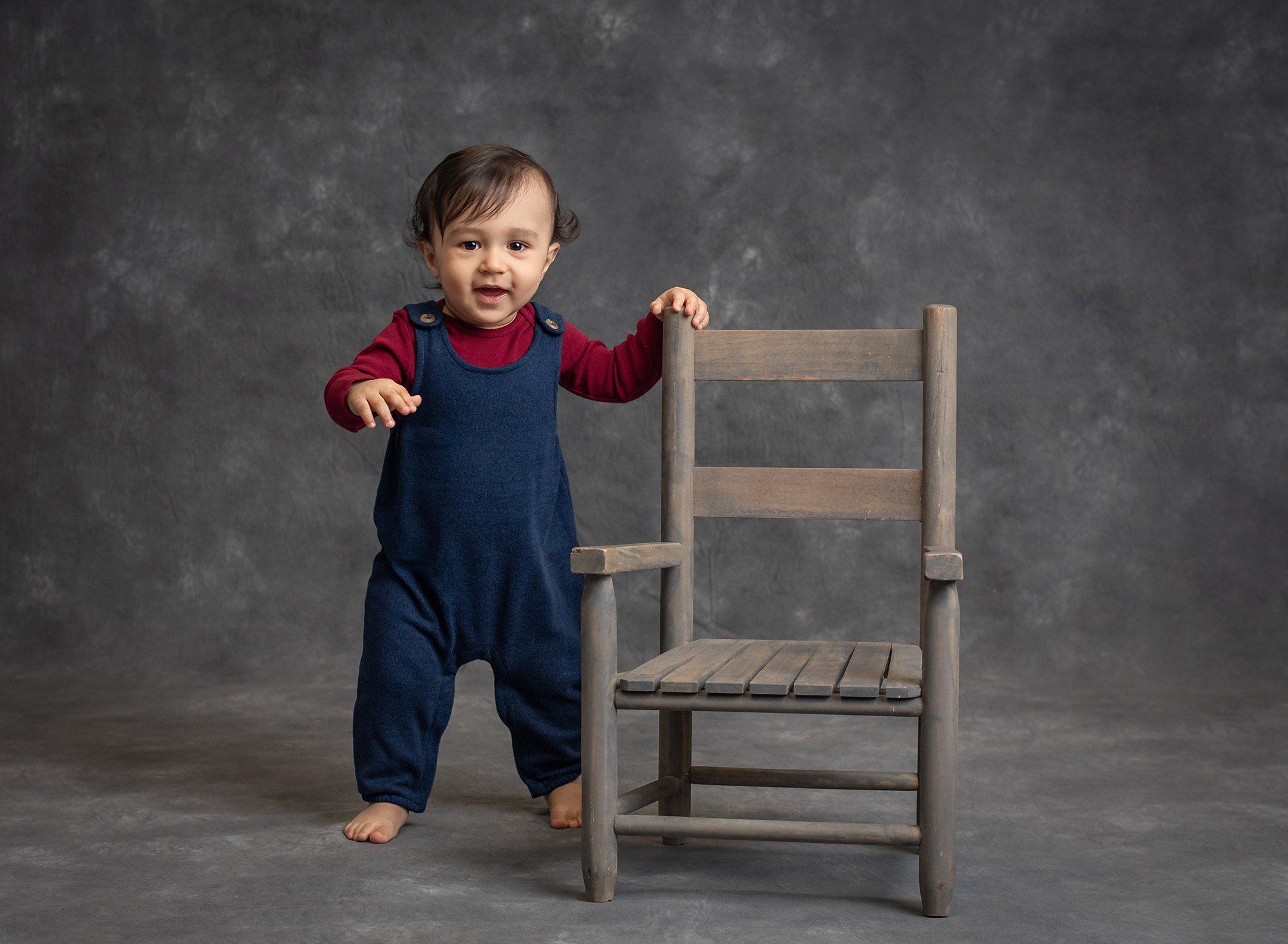 one year old boy standing with hand on rustic chair on grey background