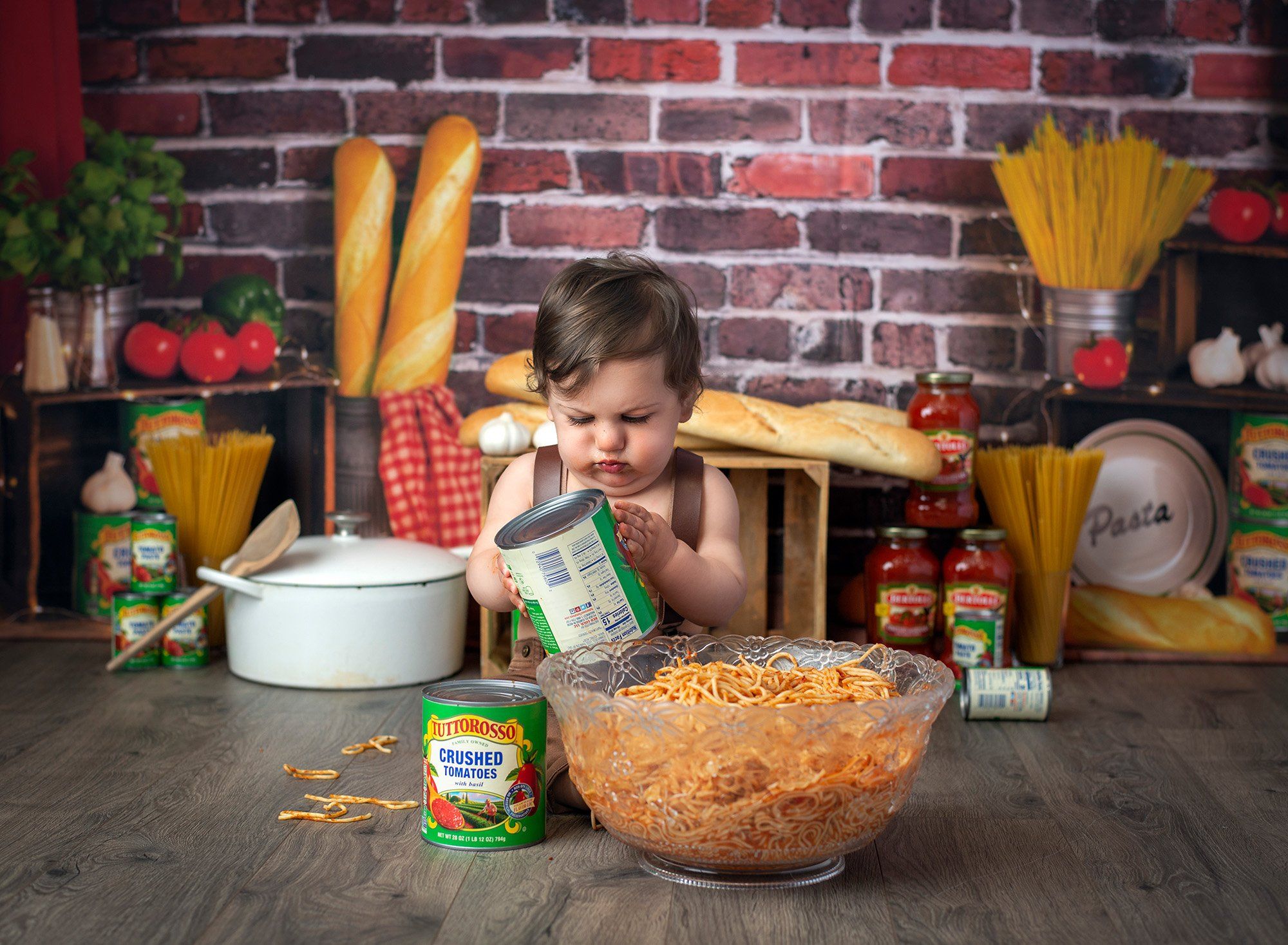 one year old boy playing with tomato cans while surrounded by pasta and sauce jars on a brick background