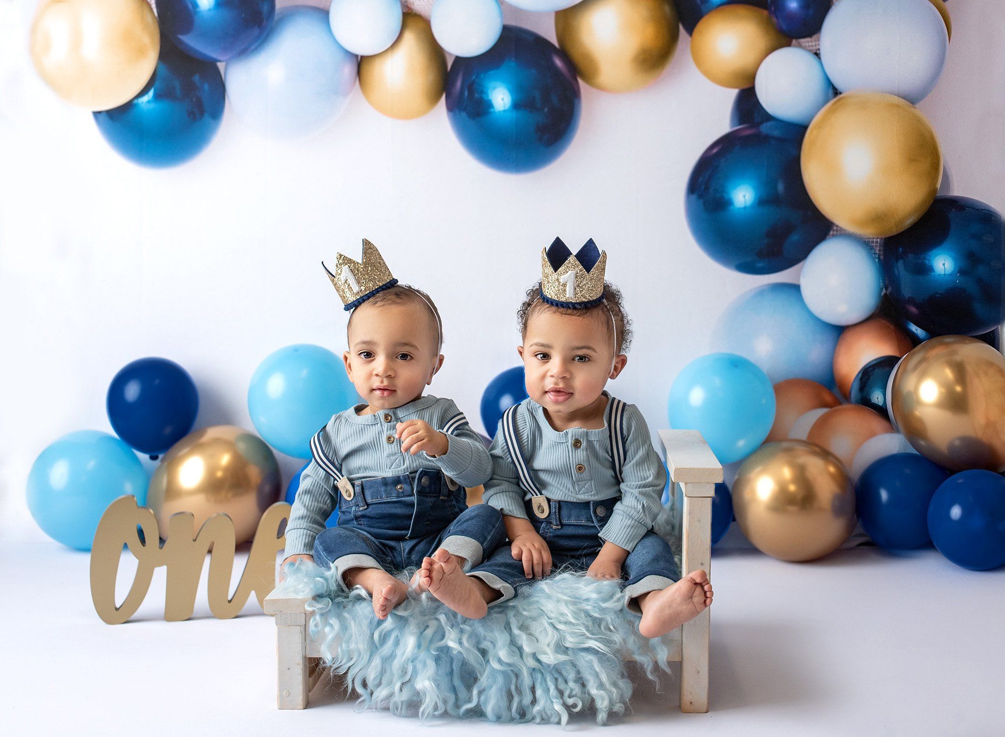 one year old twin boys sitting on white miniature bed wearing crowns with blue and gold balloons in background with one sign