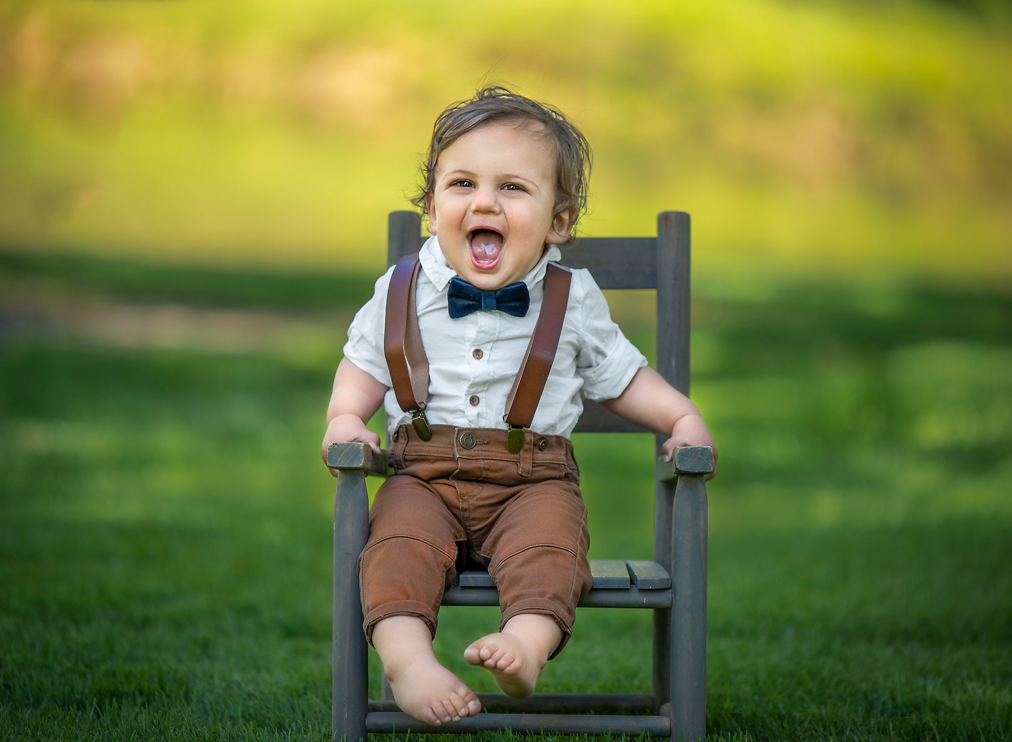 1 year baby photoshoot one year old boy wearing overalls laughing while sitting in a rustic chair