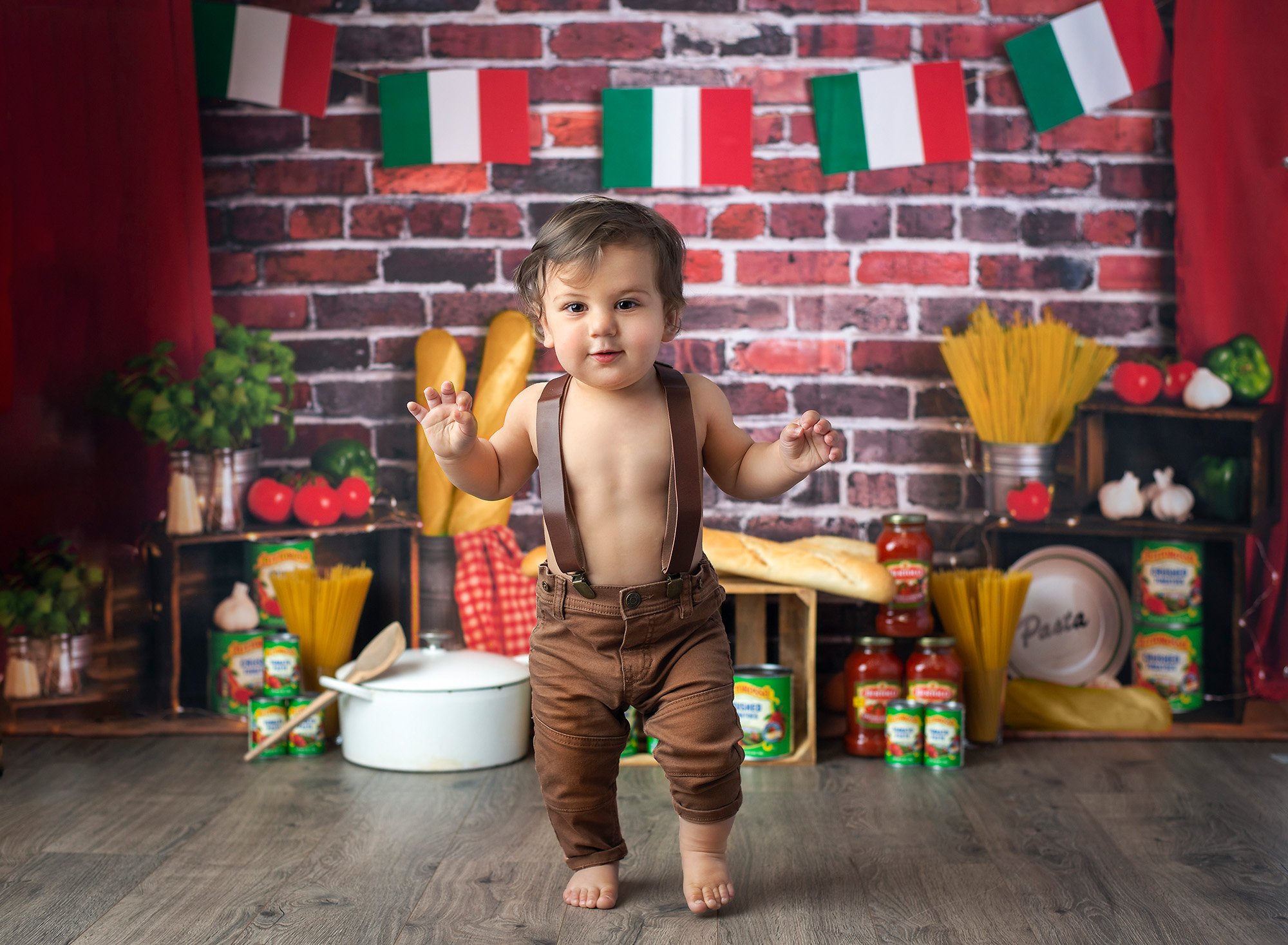 1 year baby photoshoot spaghetti smash one year old boy walking in overalls with Italian flags, Italian bread, pasta and jars of sauce in background