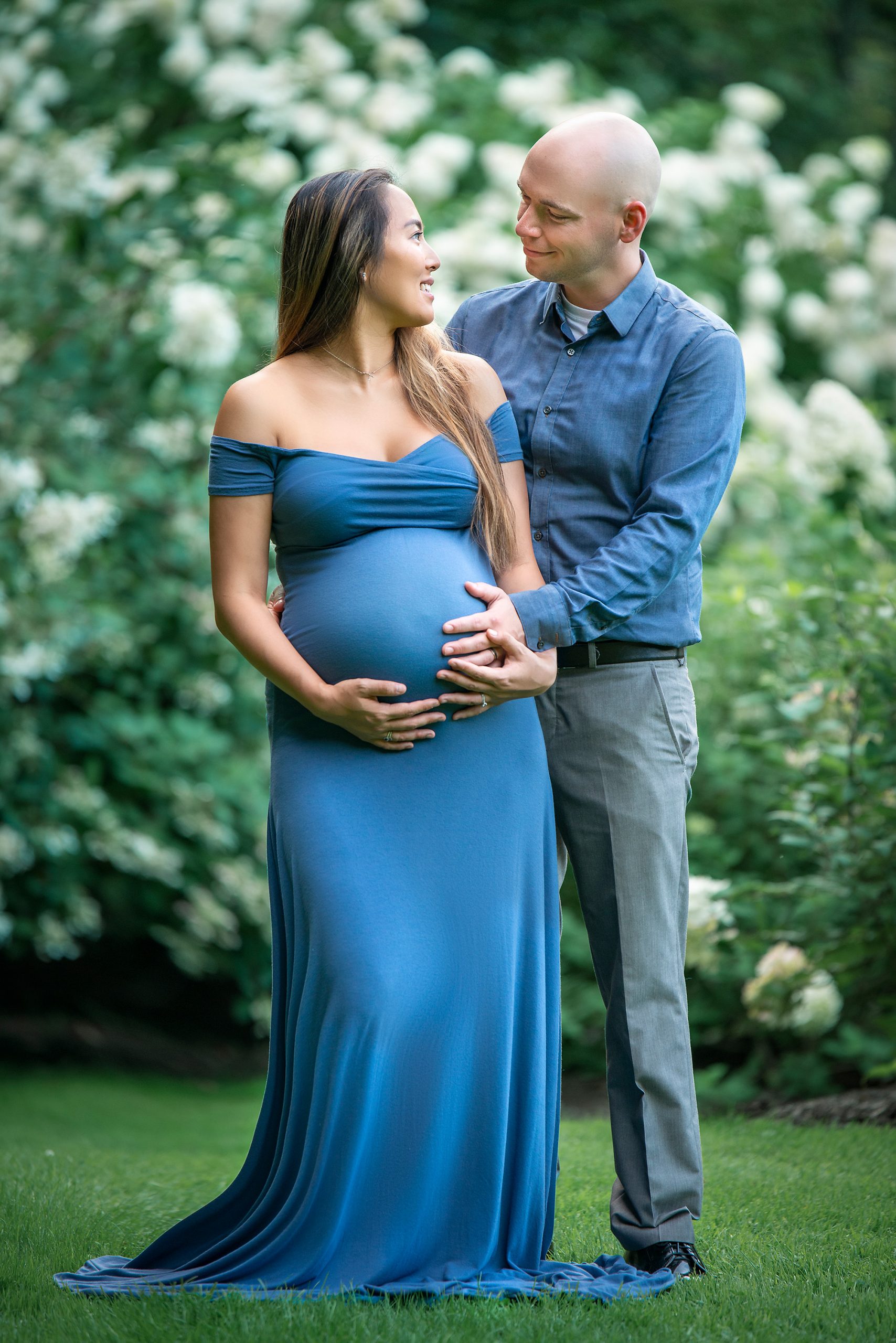 pregnant woman wearing a blue dress posing with husband in front of a white flower bush
