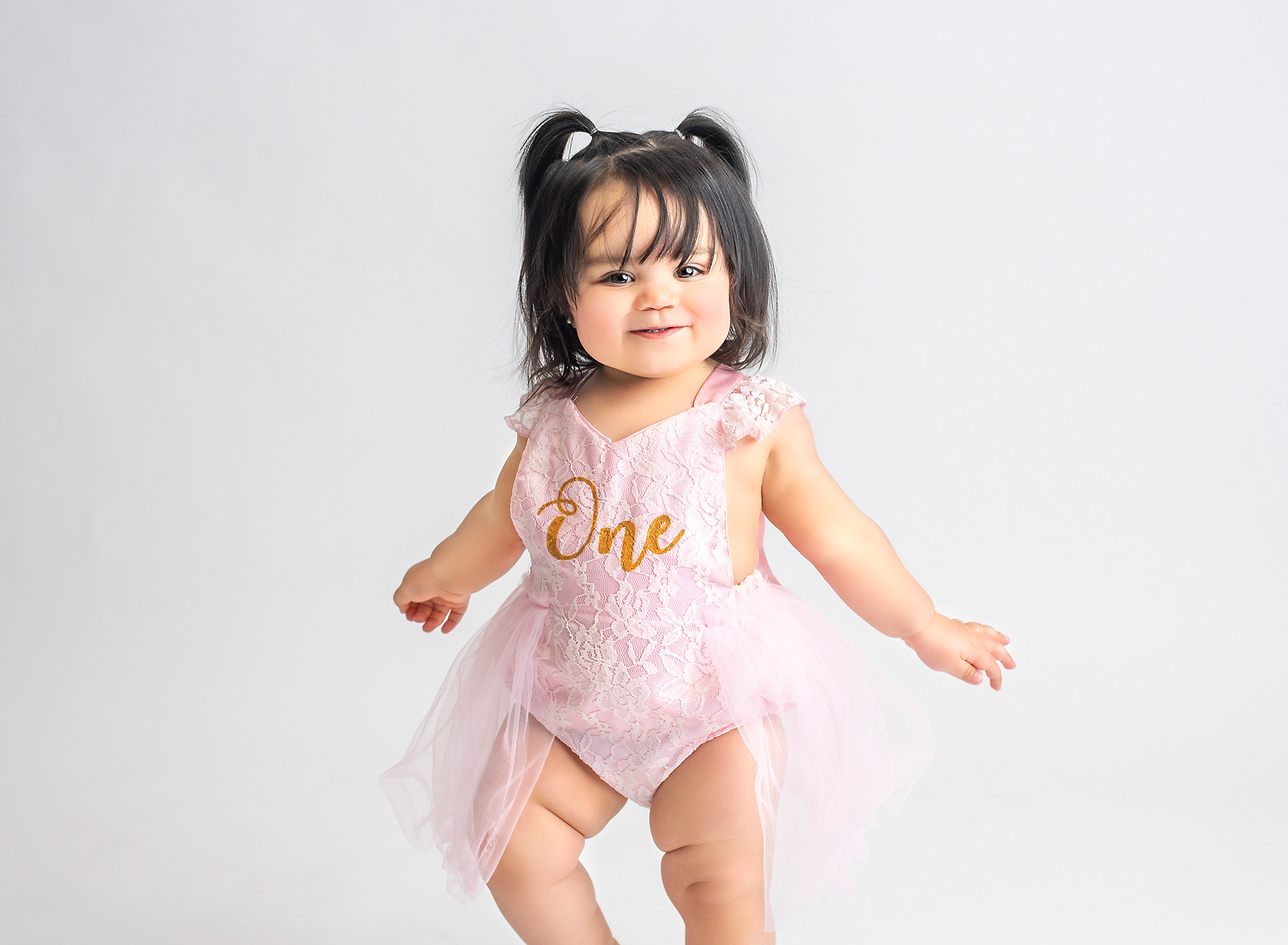 one year old baby girl in pigtails and pink laced one dress standing on neutral background
