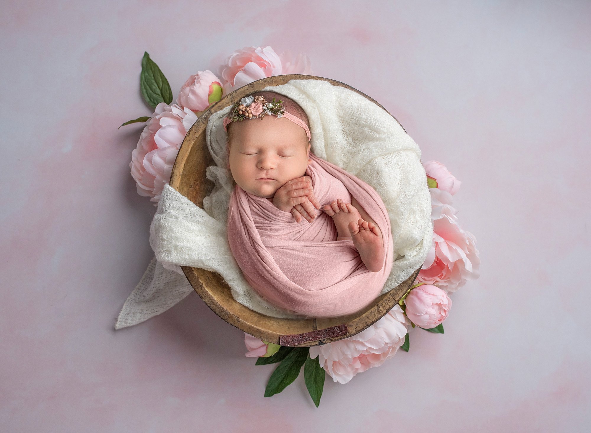 swaddled newborn pictures newborn baby girl asleep in pink wrap laying in wooden bowl surrounded by pink peonies