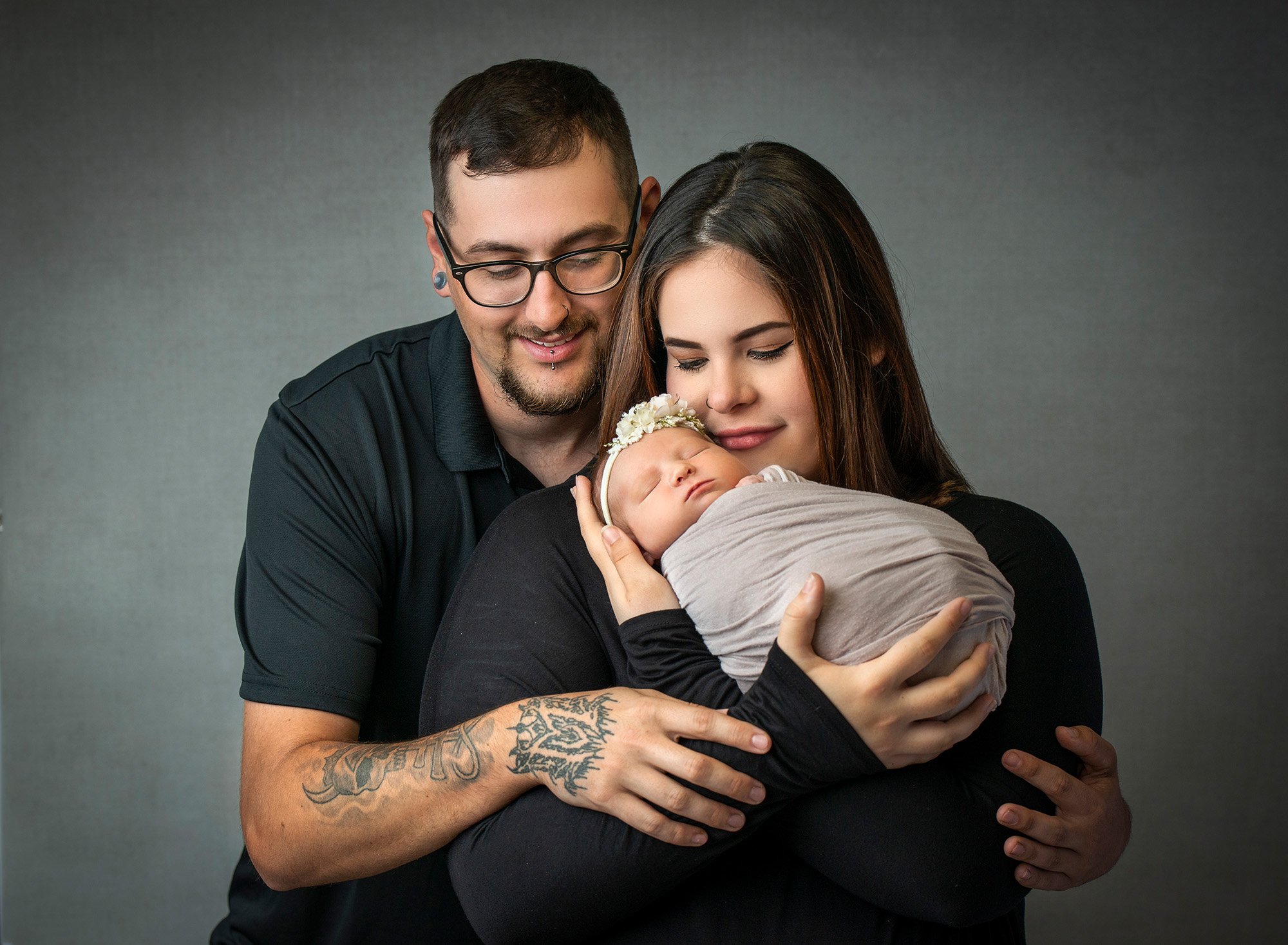 new parents cradling newborn baby girl wearing floral headband swaddled in grey wrap