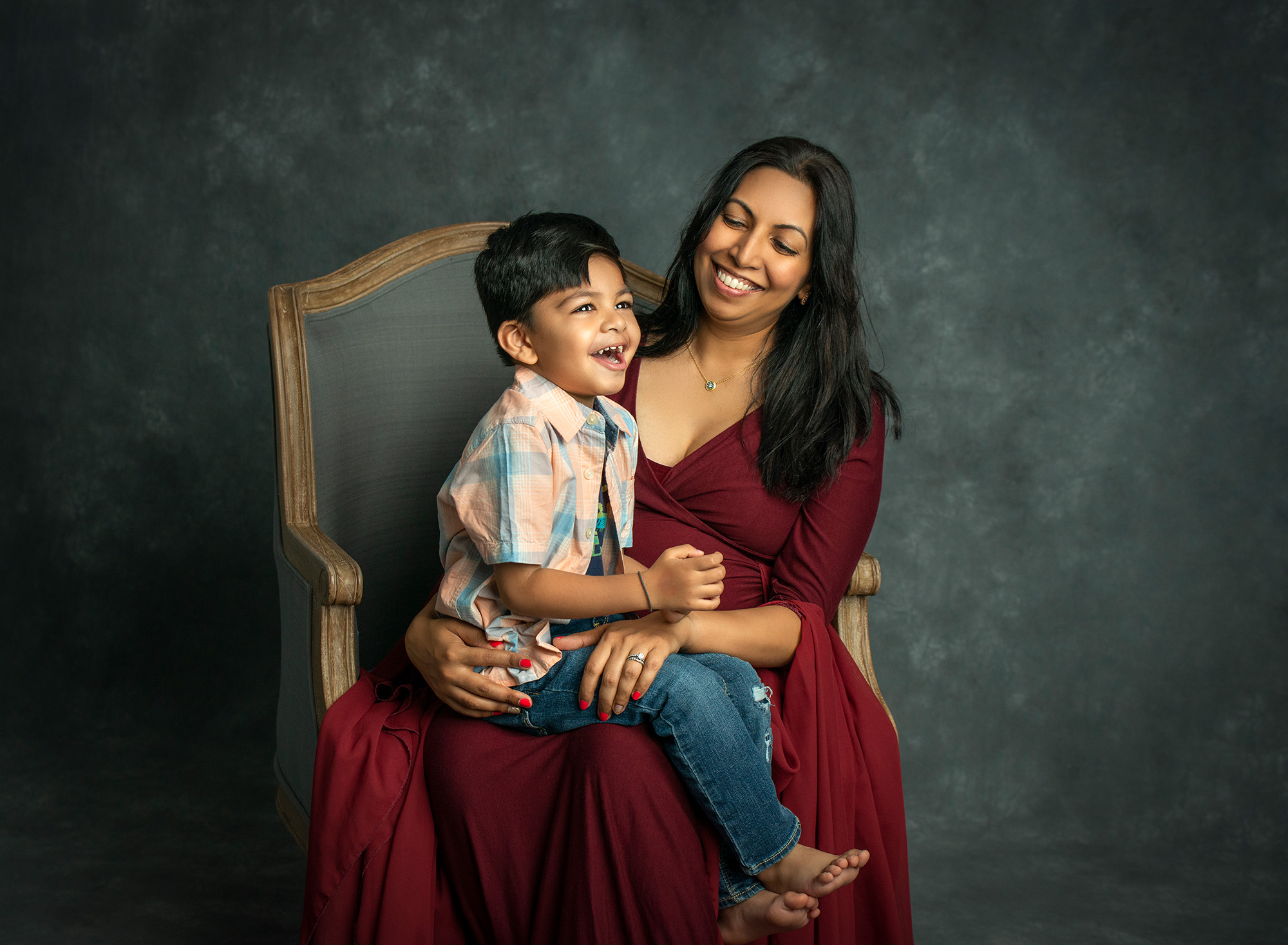 young boy smiling while sitting on the lap of his pregnant mother in red maternity gown