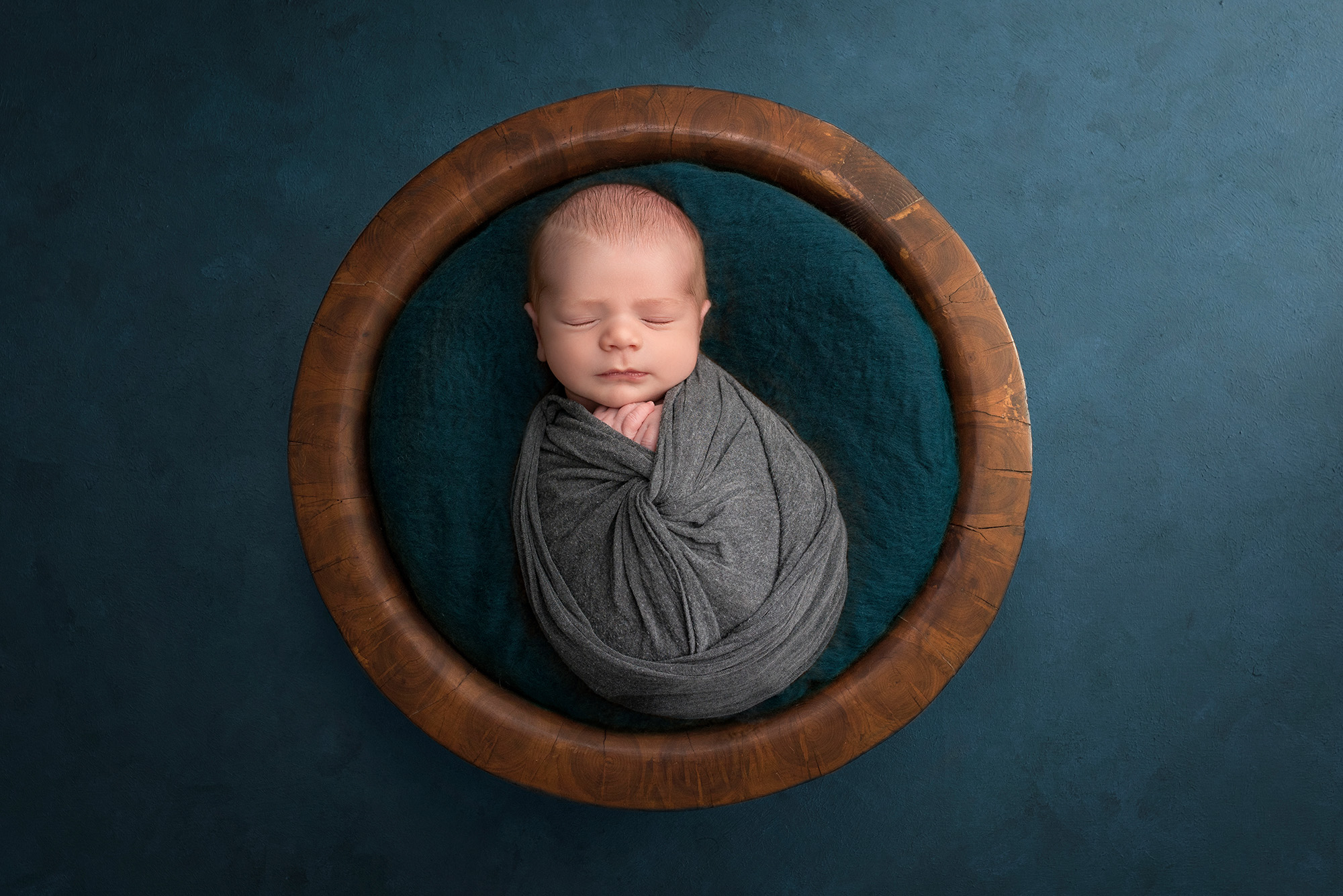 newborn baby boy asleep in wooden bowl on teal background