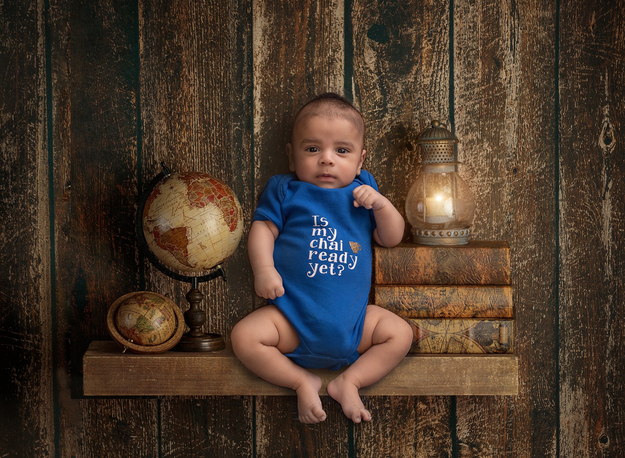 newborn boy sitting on shelf with old books, world globe maps and a lantern on a wooden backdrop