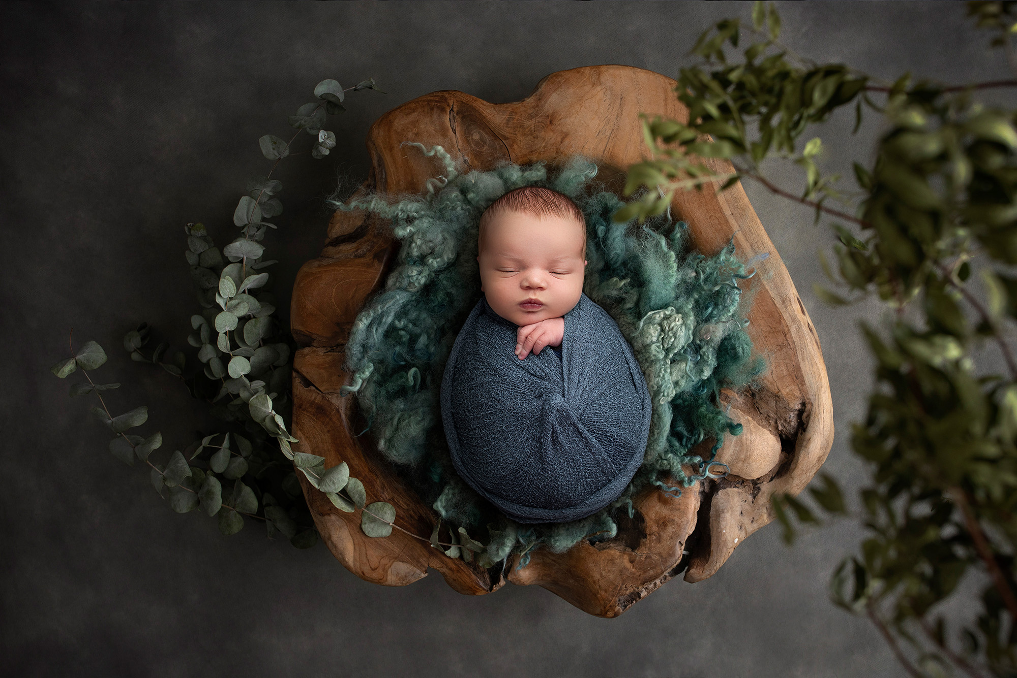 newborn baby boy asleep on wooden bowl surrounded by greenery