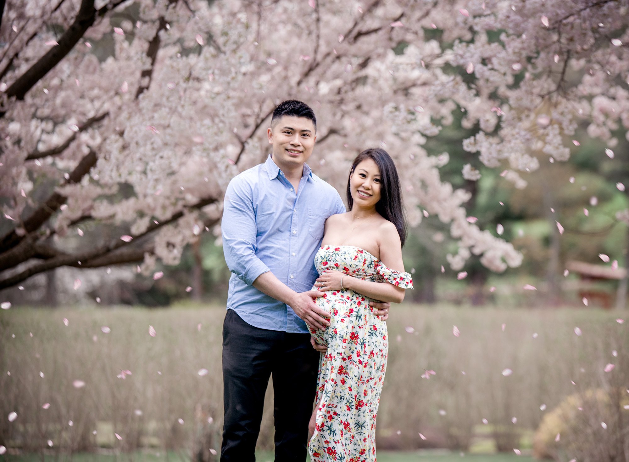 Spring Maternity Photos in Manchester CT pregnant woman in floral maternity dress posing with partner under a weeping cherry tree while petals are falling