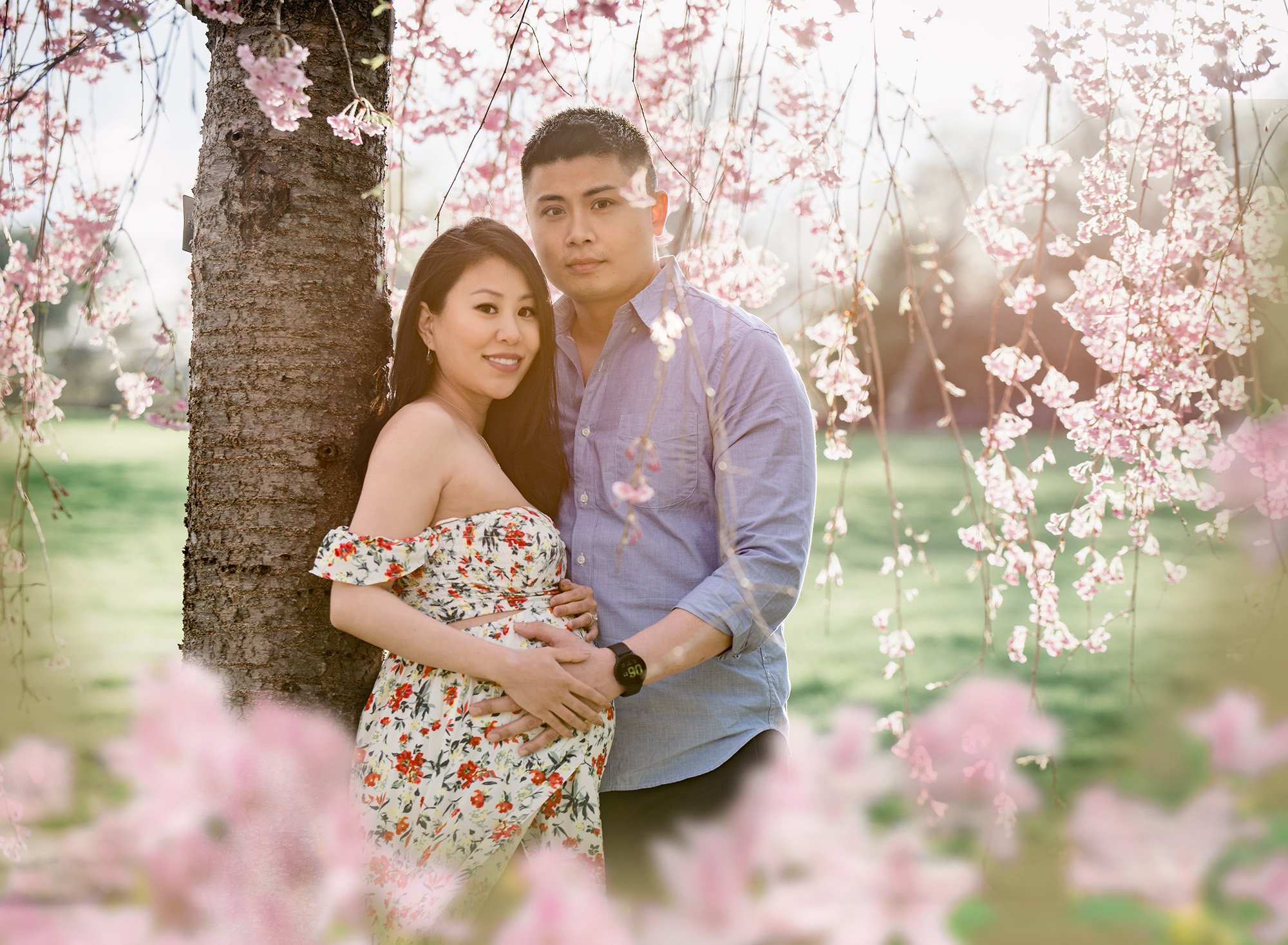 garden flower maternity photos pregnant woman in floral maternity dress posing with partner under a weeping cherry tree