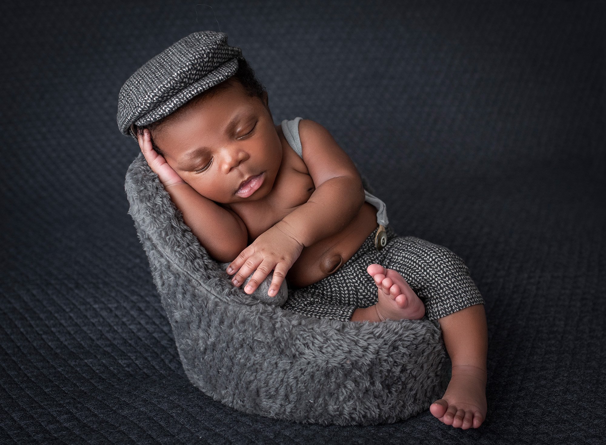 Jamaican boy newborn photographs newborn baby boy asleep in gray lounge chair wearing Gatsby cap