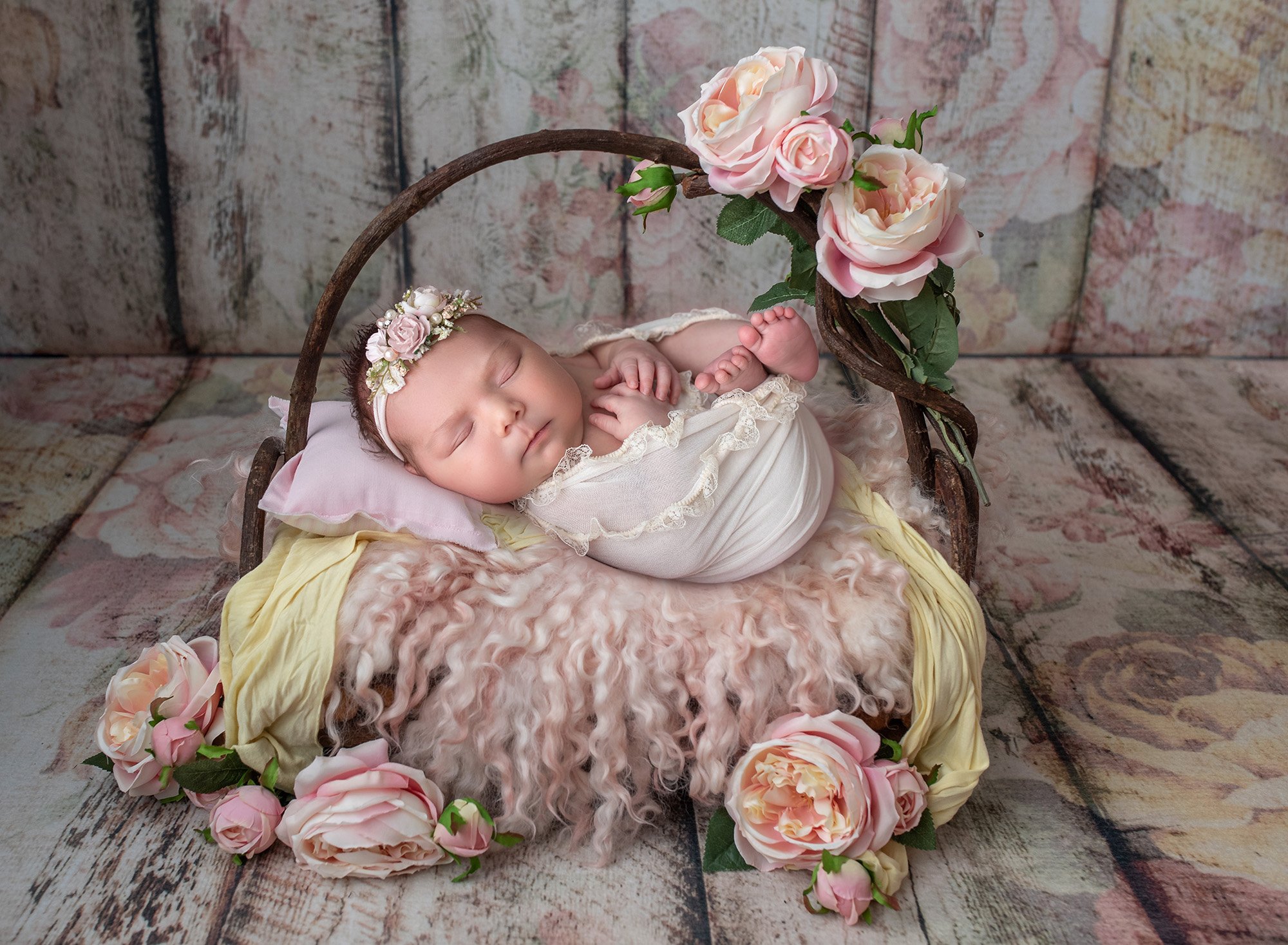 newborn baby girl swaddled in lace wrap sleeping on top of rustic wooden bed with pink fuzzy blanket surrounded by pink flowers on floral background