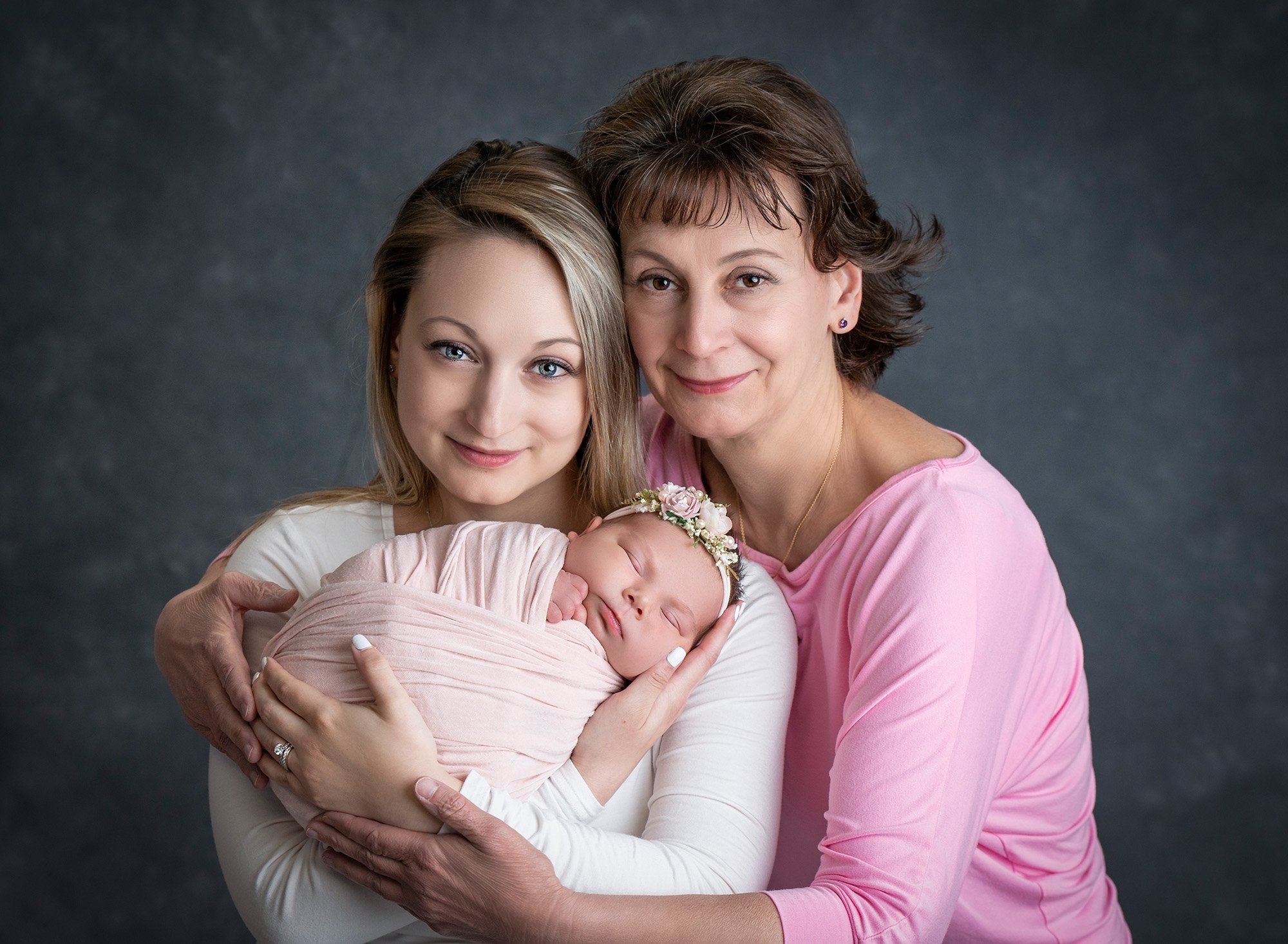 mother and daughter holding newborn baby girl swaddled in soft pink wearing floral headband on dark gray backdrop