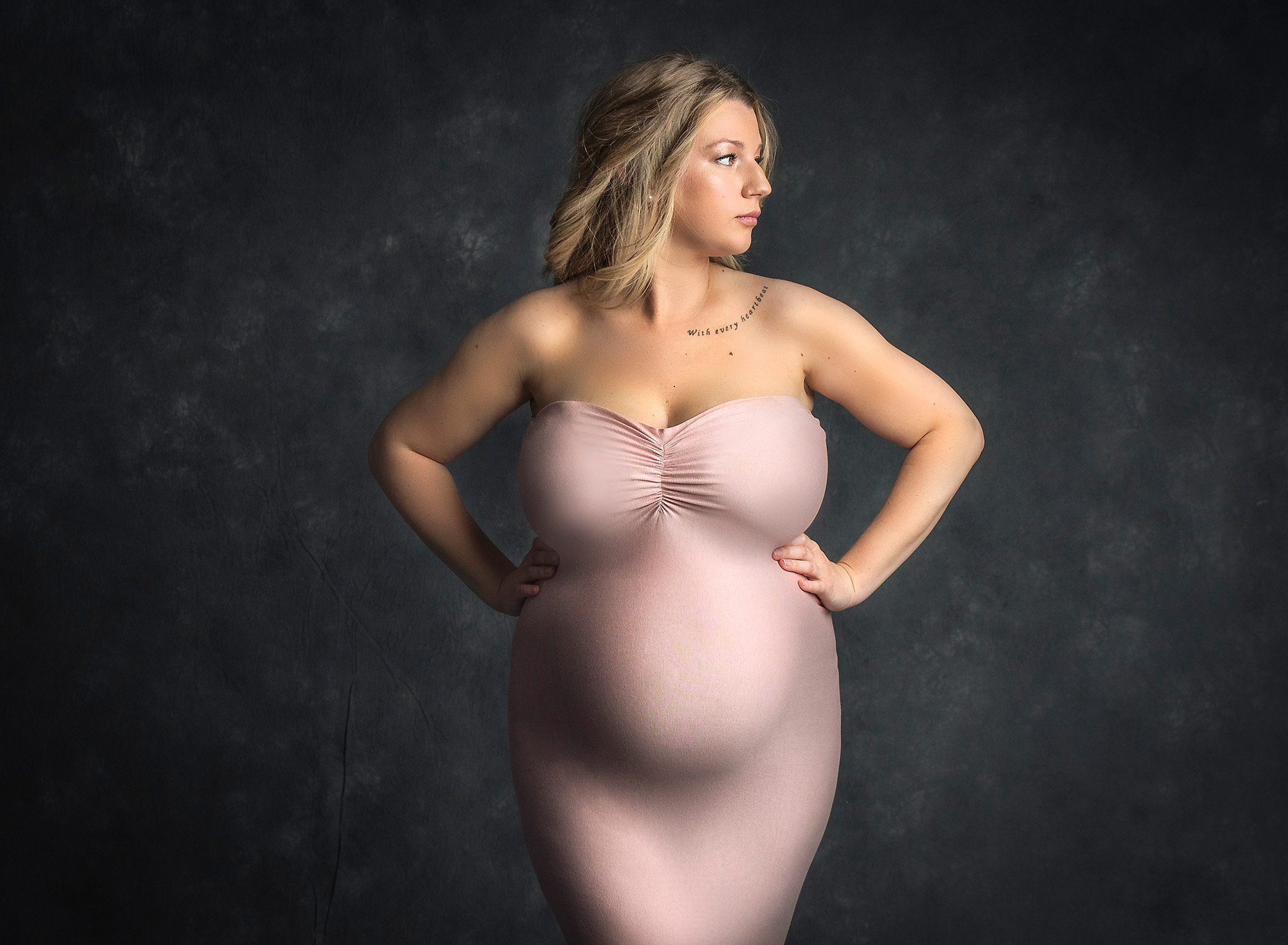 blonde pregnant woman posing in pink strapless maternity dress on dark grey background