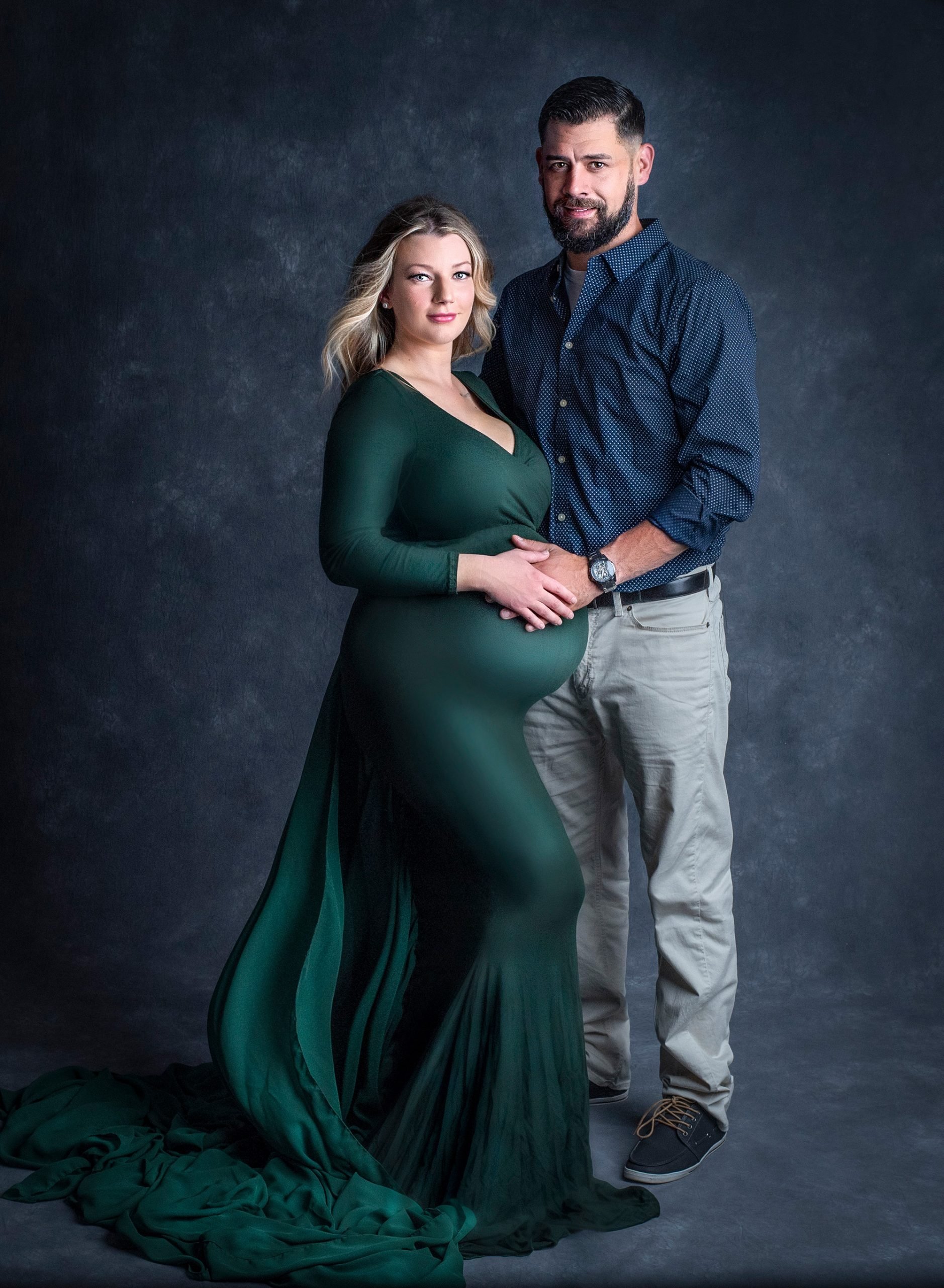 Studio Maternity Photos Glastonbury CT blonde pregnant woman posing in green maternity dress while holding hands with her partner on her stomach