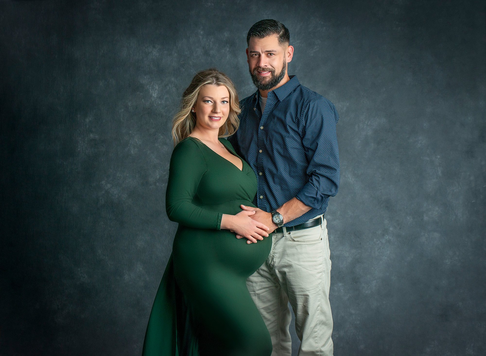 blonde pregnant woman in green maternity dress posing with partner holding hands on her stomach