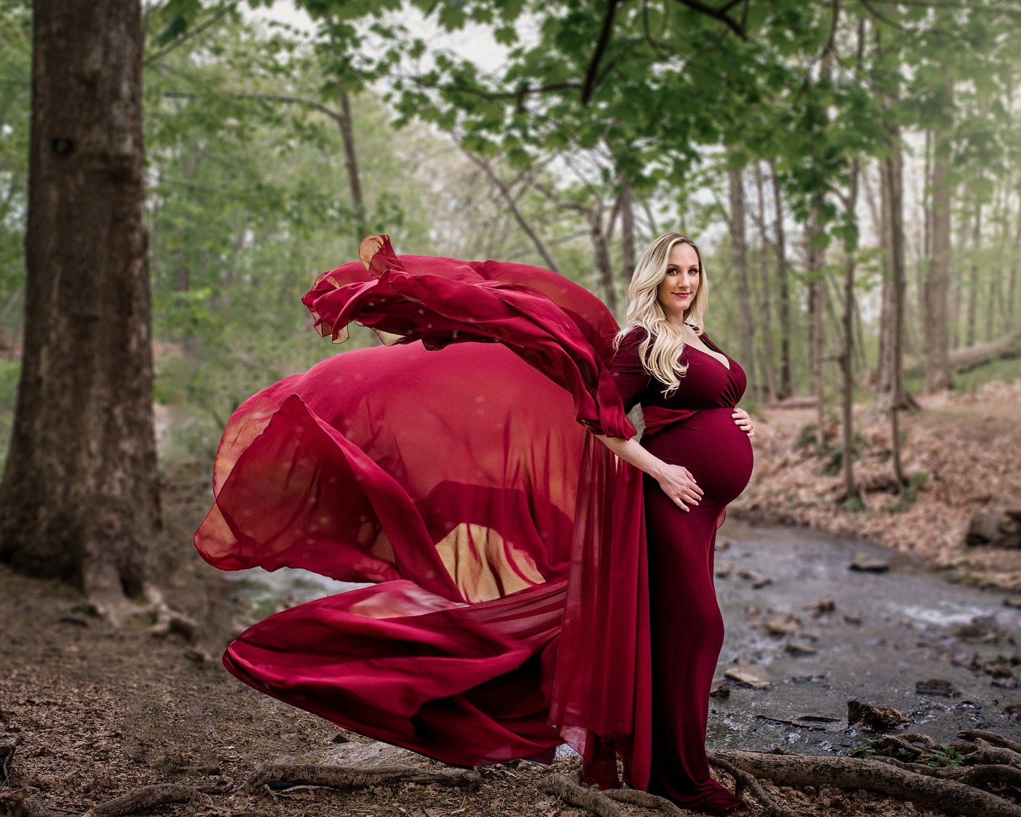 Connecticut Maternity Photographer Beautiful blonde pregnant woman wearing a flowing red gown in front of a river in the woods