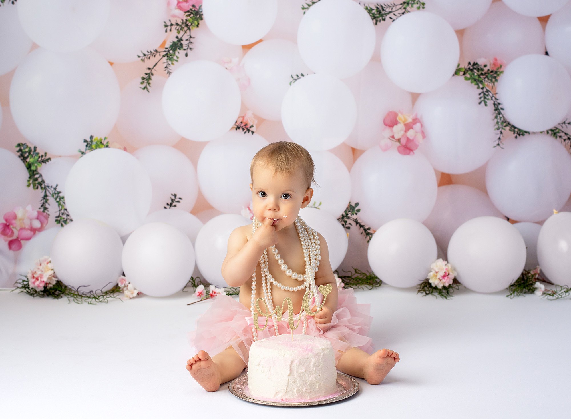 baby girl wearing layered pearl necklace and pink tutu sitting in front of cake with gold glitter ONE sign with white balloons in background putting finger in mouth