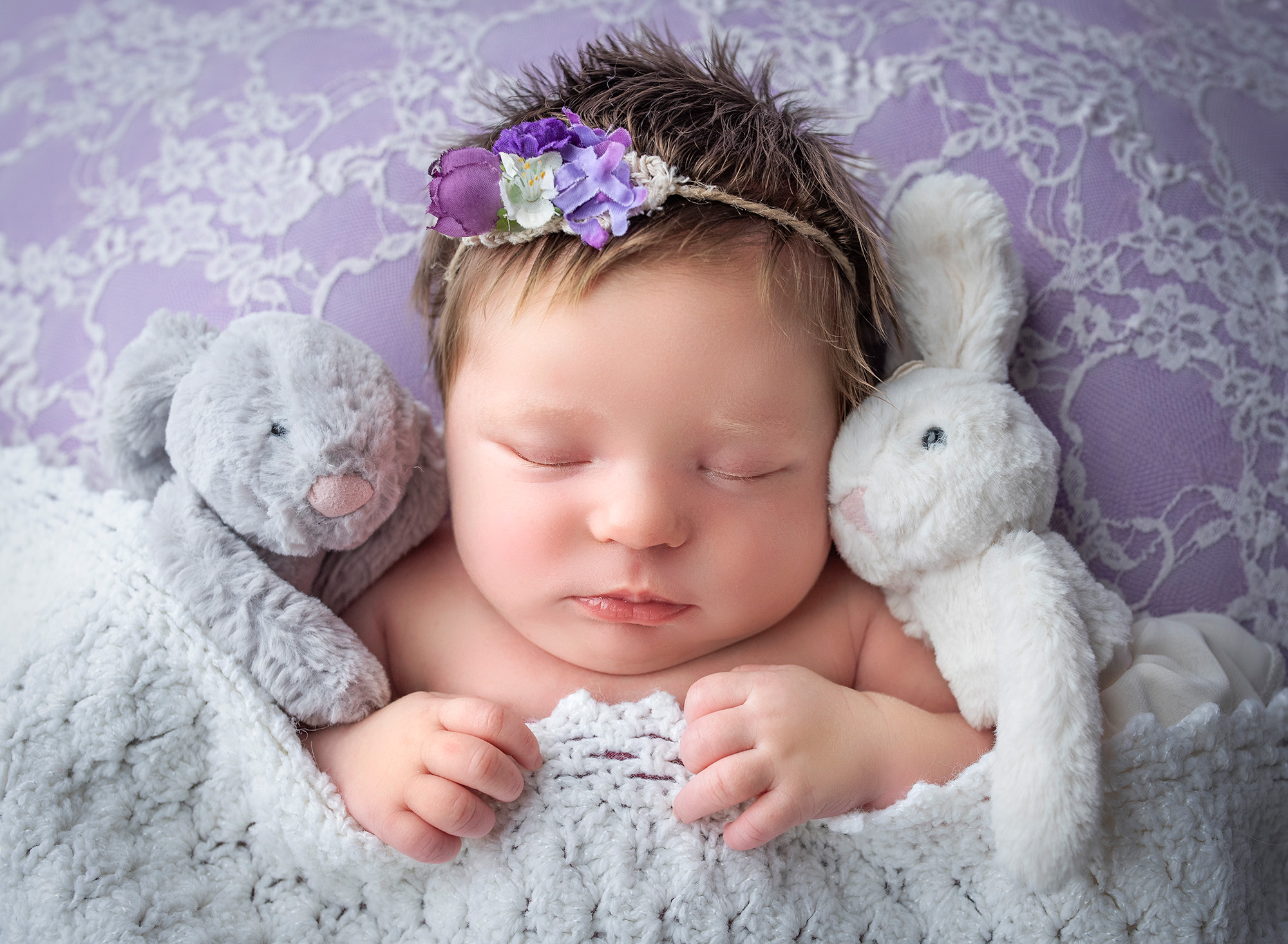 newborn baby girl asleep under blanket wearing purple headband with stuffed bunnies on each side of her