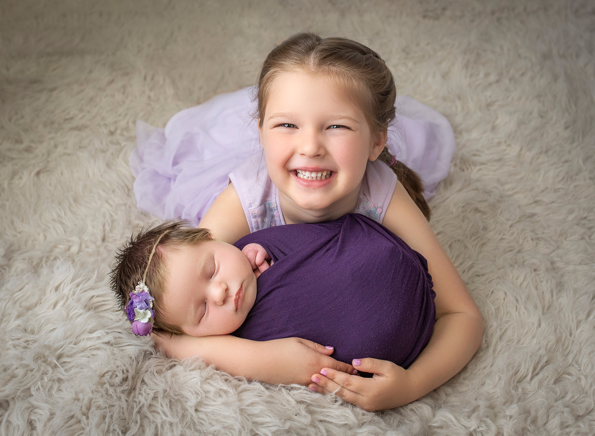 Newborn Sibling Photos smiling young girl wearing purple dress cradling newborn baby sister swaddled in purple