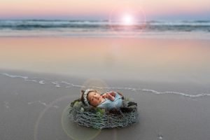 newborn baby girl laying in a basket on the sand at the beach while the sun is setting