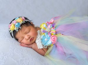 newborn baby girl asleep in a multi color floral costume on laced blanket