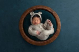 newborn baby dressed in bear sweater romper asleep in wooden bowl