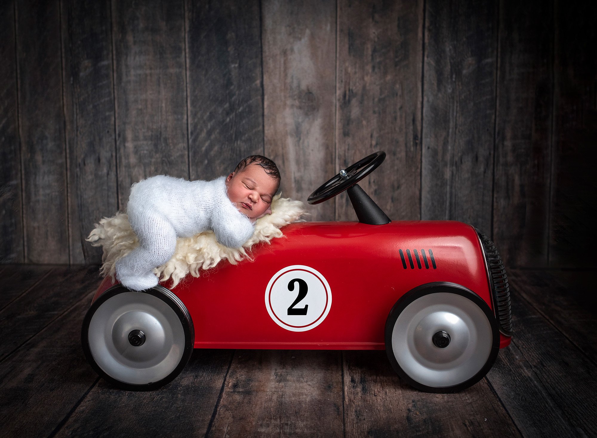 newborn baby boy in sweater romper asleep on a red car