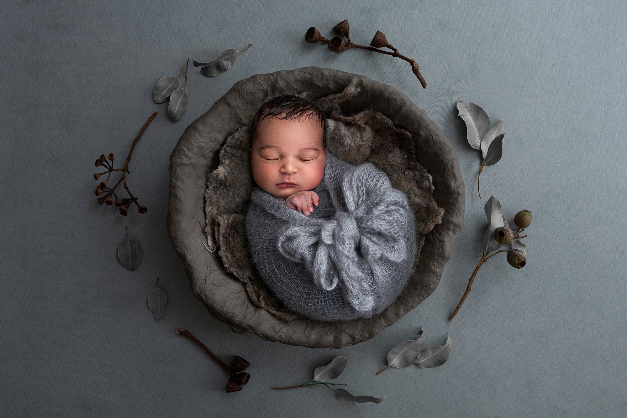 newborn baby asleep in a bowl swaddled in a blue blanket surrounded by dried flowers