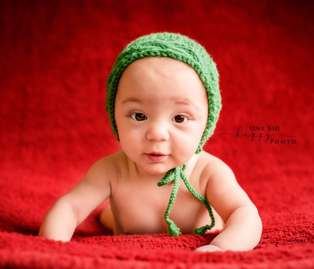 Baby lying on his tummy pushed up on a red blanket with a bright green hat on