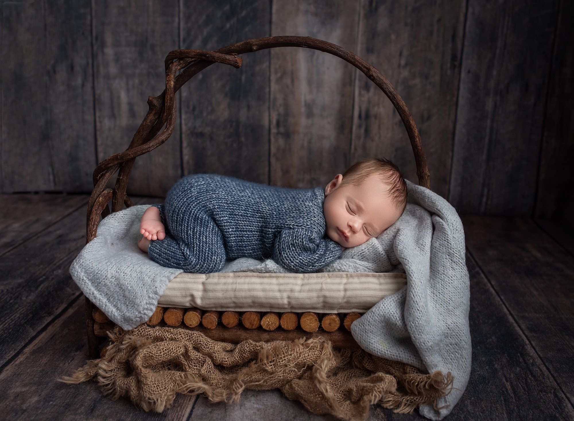 newborn baby boy asleep in wooden basket wearing blue sweater romper on wood background