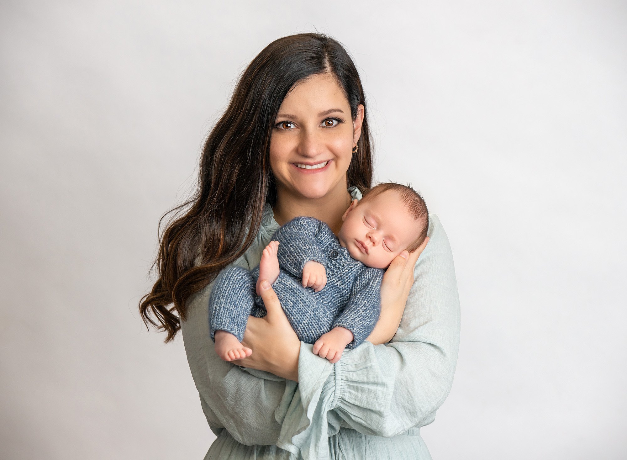 new mom holding newborn baby boy wearing blue sweater romper on white background