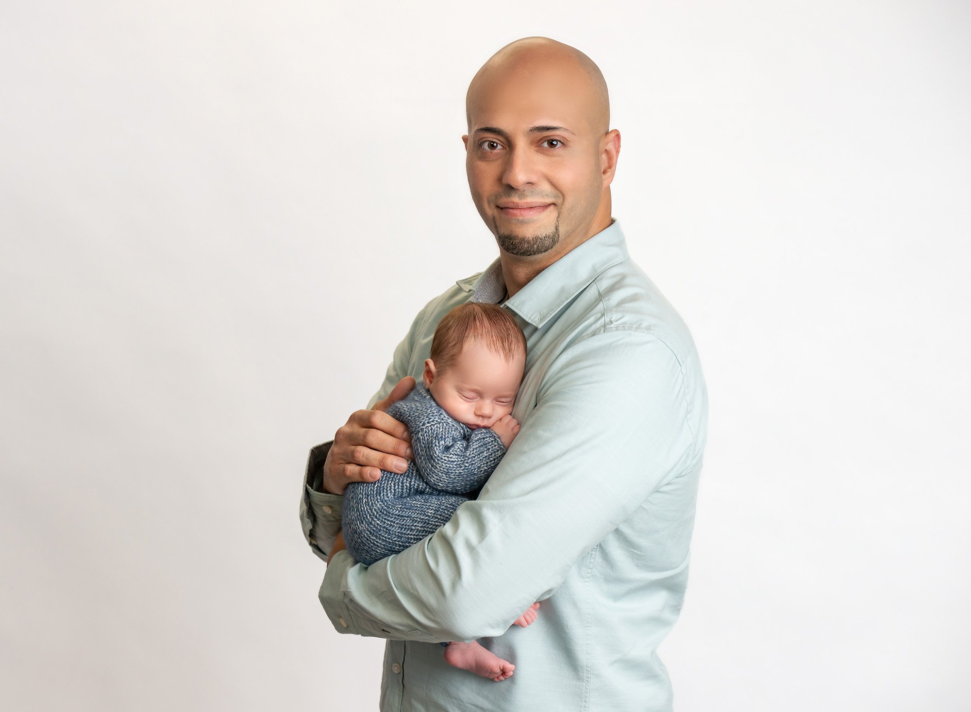 new dad holding newborn baby boy wearing blue sweater romper on white background