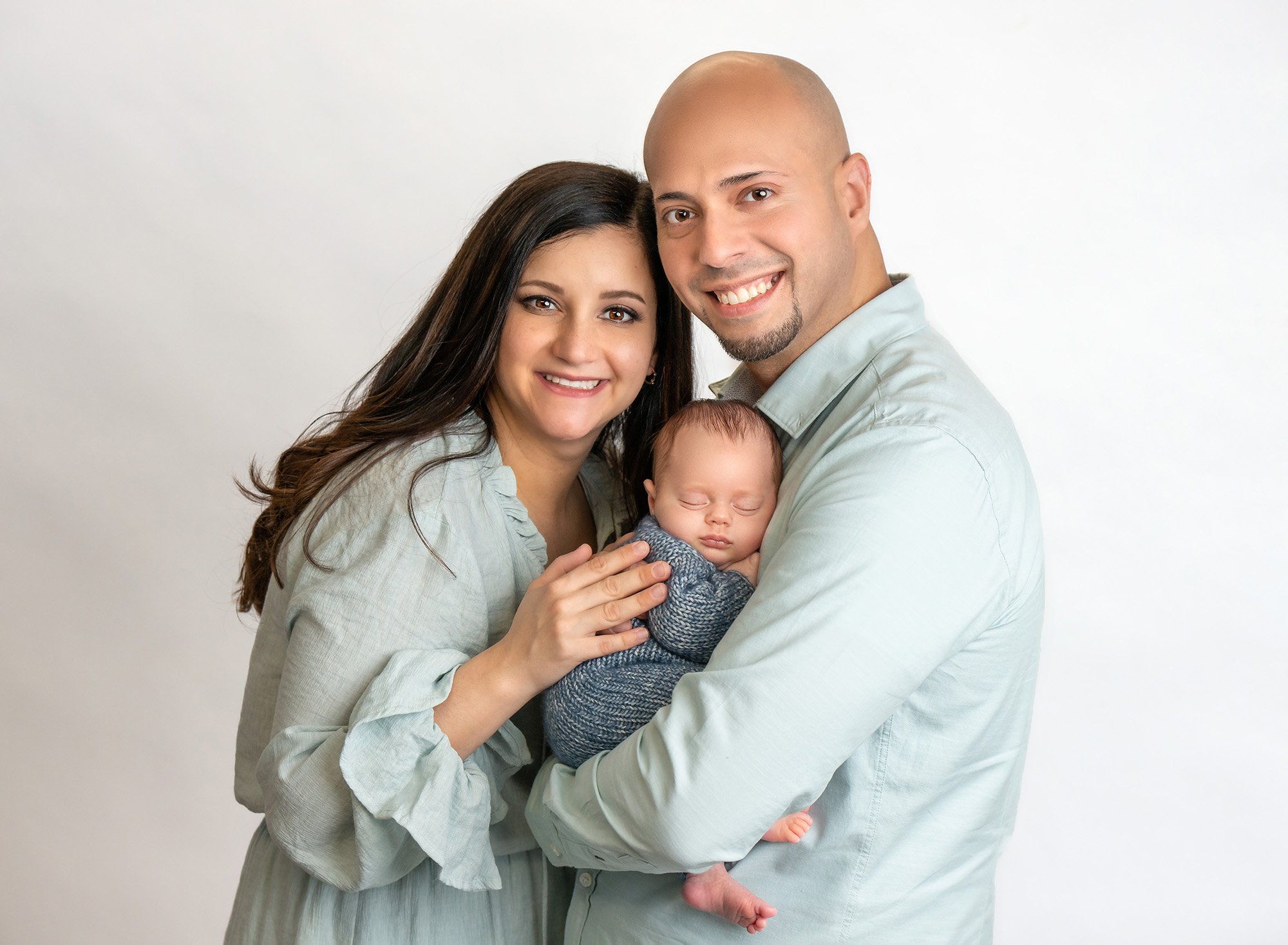 new parents dressed in light blue holding newborn baby boy in blue sweater on white background