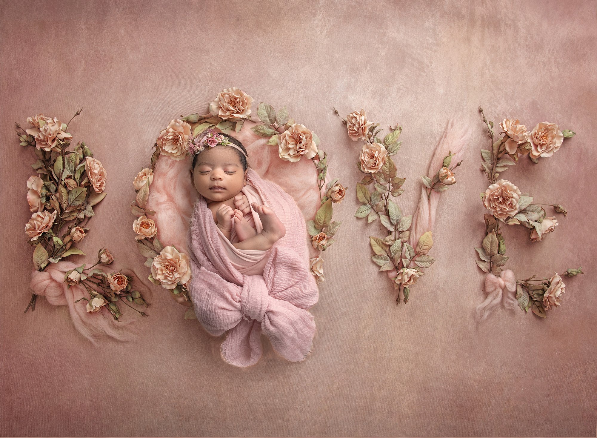 sweet newborn baby girl swaddled in pink wrap asleep on a pink background spelling out the word LOVE in flowers and bows