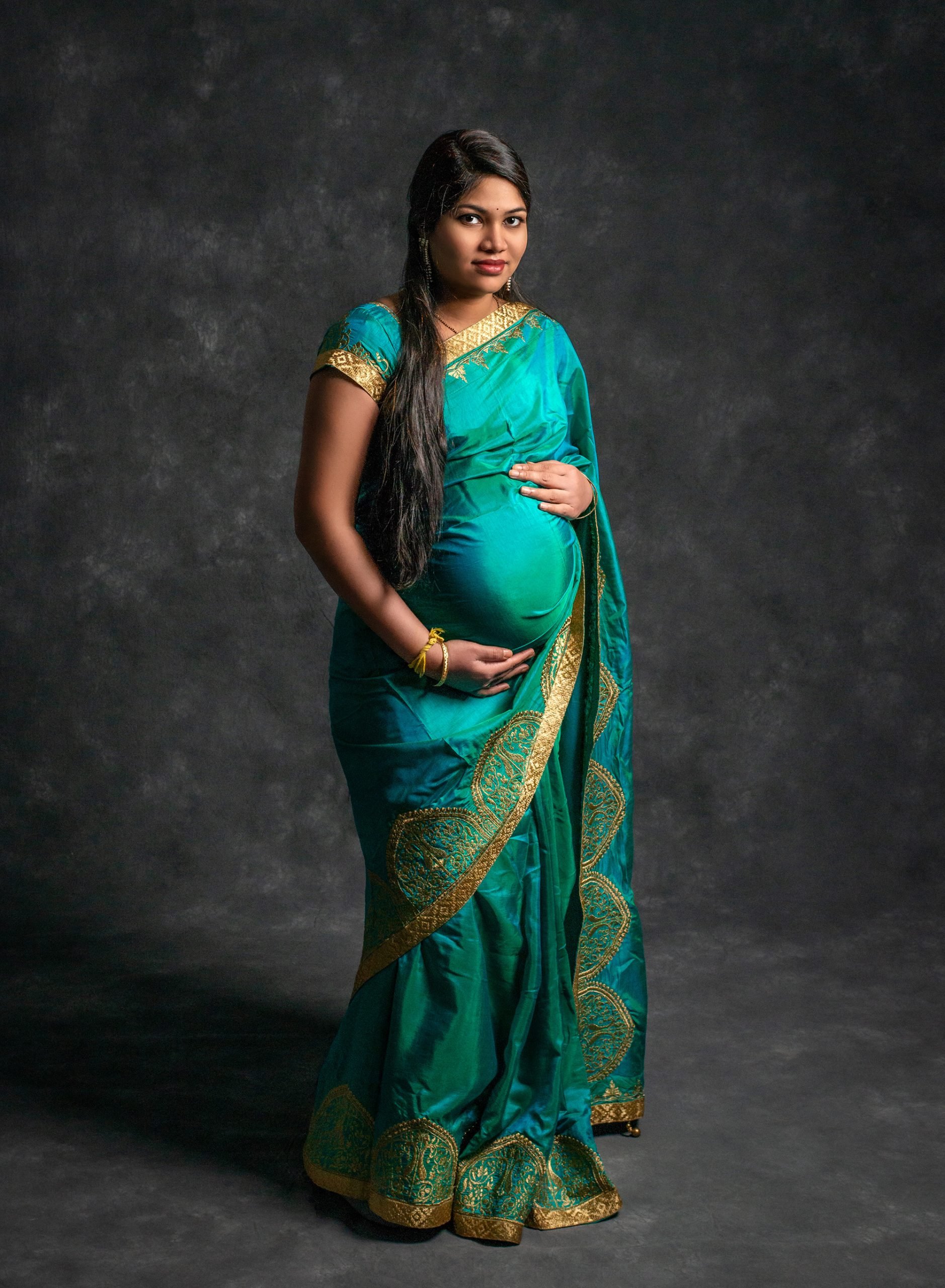 Traditional Indian clothes maternity photo pregnant woman posing holding stomach in Indian dress clothing on gray backdrop