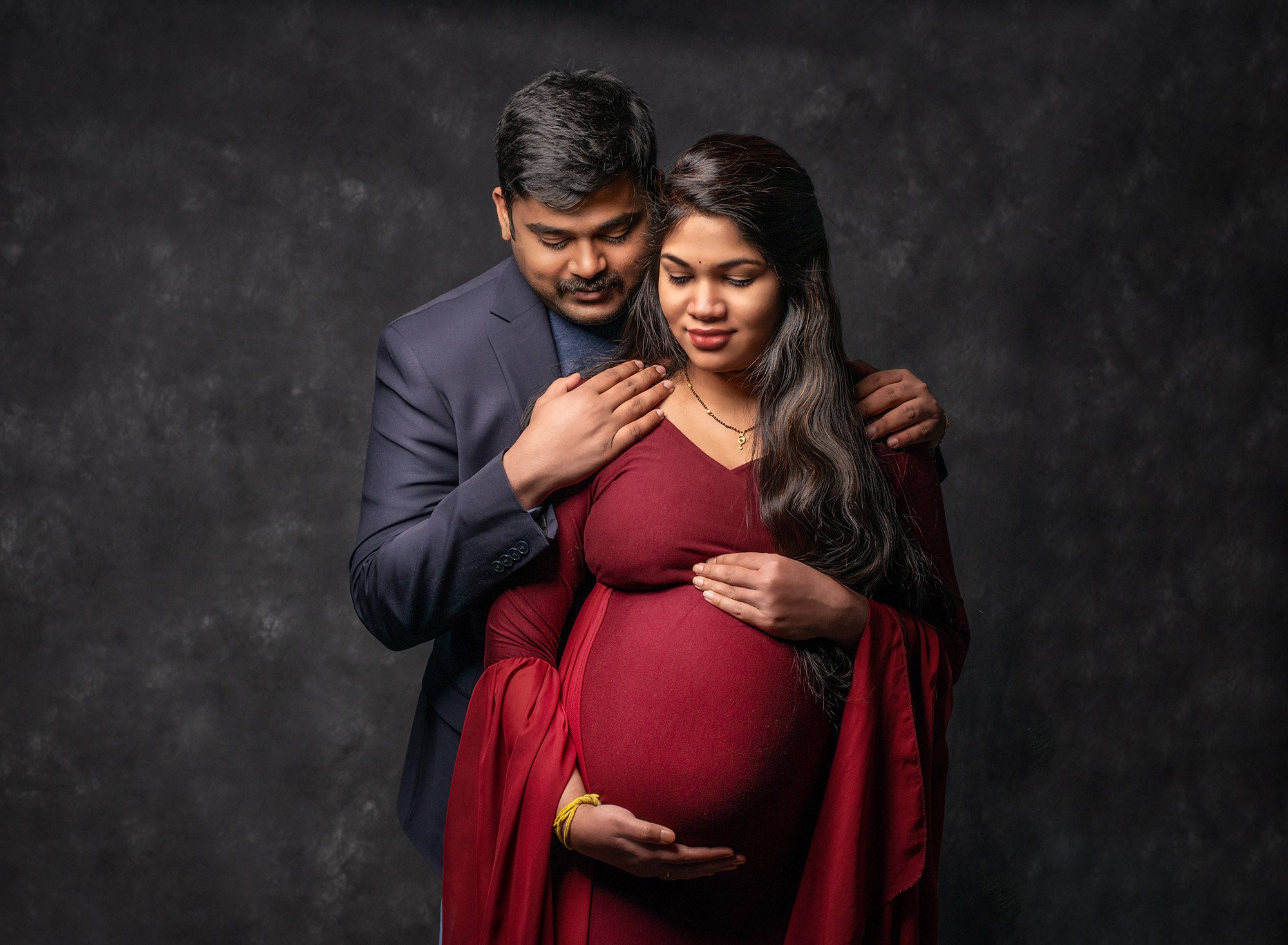 husband looking down at wife's pregnant stomach in cranberry maternity gown