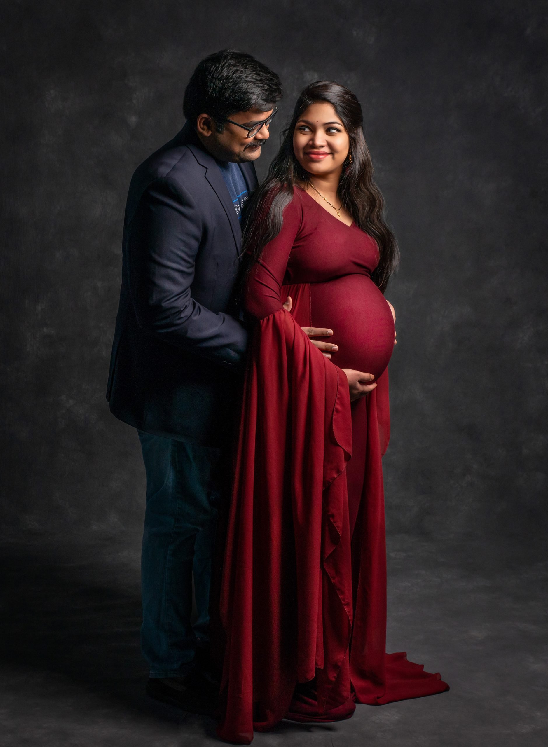 pregnant woman in cranberry maternity gown while partner holds her stomach