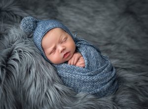 newborn baby boy swaddled in blue sweater blanket and bonnet atop a fluffy blanket
